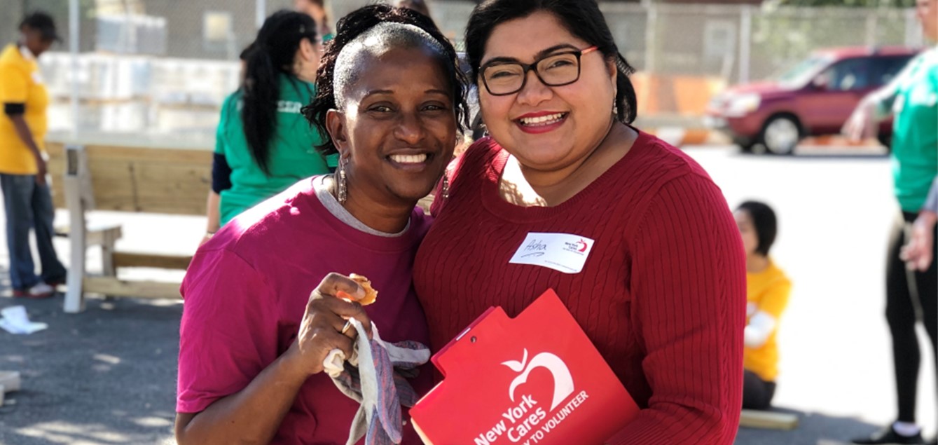 Two women smiling wearing red shirts and holding New York Fares swag