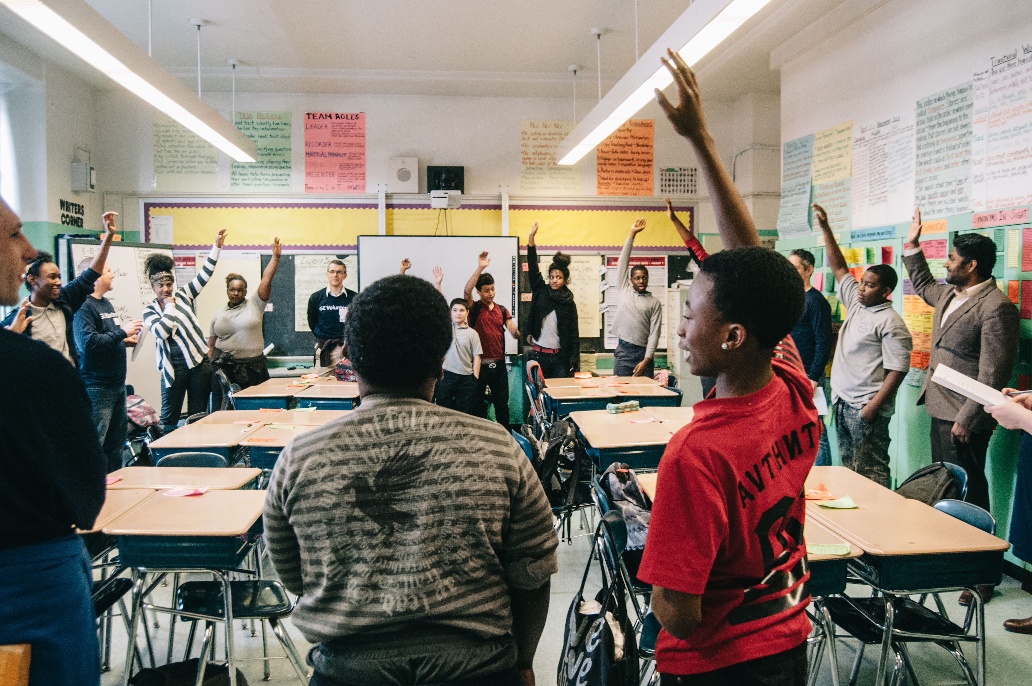 18 students stand in a circle around classroom desks and raise their hands excitedly